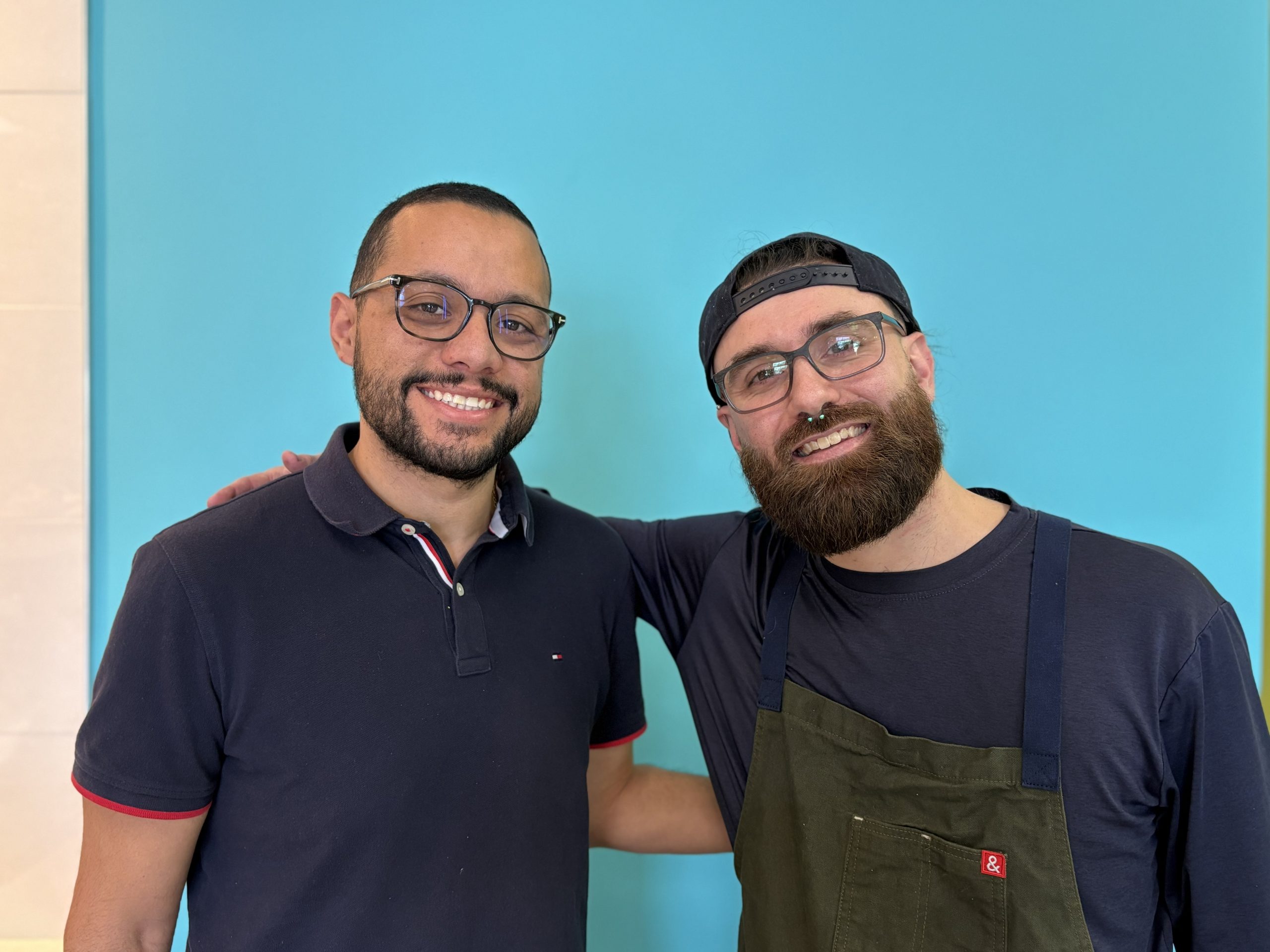 Shawn Mello and Abe Agayby, co-owners of Farm2Cup Fairhaven, smiling together in a vibrant cafe setting with a blue background, emphasizing community and friendship in their local business.